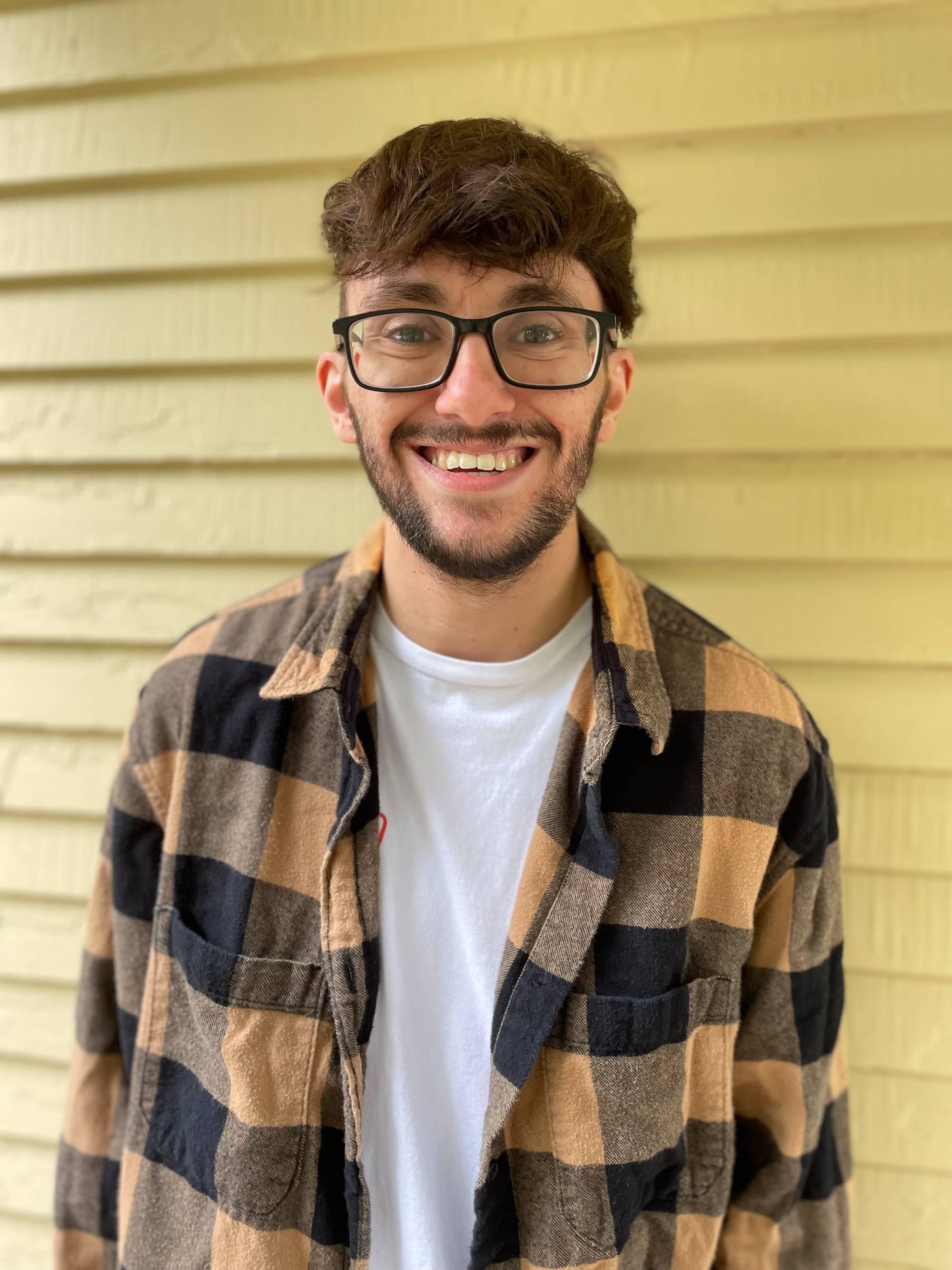 Smiling man with brown hair and beard wearing flannel shirt with yellow background